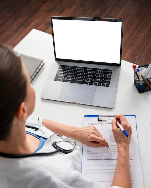 A person sitting at a desk, thoughtfully reviewing medical records and insurance documents, with a laptop displaying information about health insurance plans in the background.