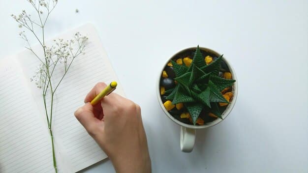 A hand holding a pen over an open journal, with a small plant and a cup of tea in the background, conveying a sense of quiet contemplation.