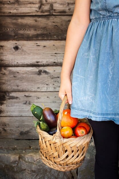 A person's hands holding a grocery basket overflowing with fresh, colorful vegetables from a farmer's market, focusing on the abundance and freshness of the produce.