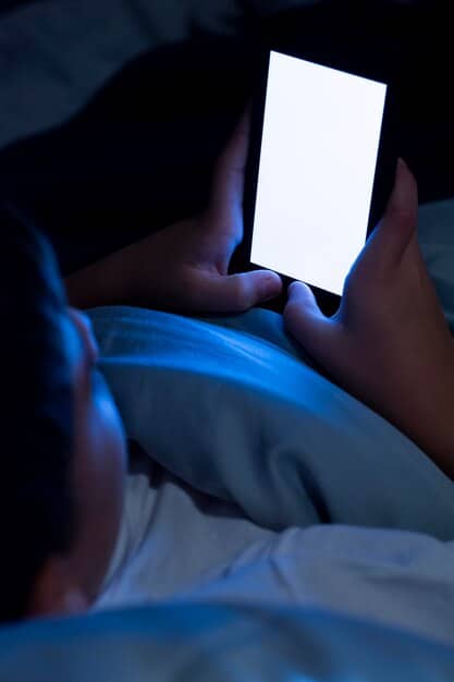A person lying in bed at night, illuminated by the bluish glow of a smartphone screen, with a clock on the nightstand showing late hours, symbolizing the interference of technology with sleep.