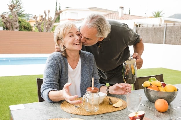 A photograph of an elderly couple enjoying a meal together outdoors. The food on the table includes various Mediterranean staples such as grilled fish, whole grain bread, vegetables, and a bottle of olive oil. The couple is smiling and engaged in conversation, emphasizing the social aspect of the Mediterranean diet.